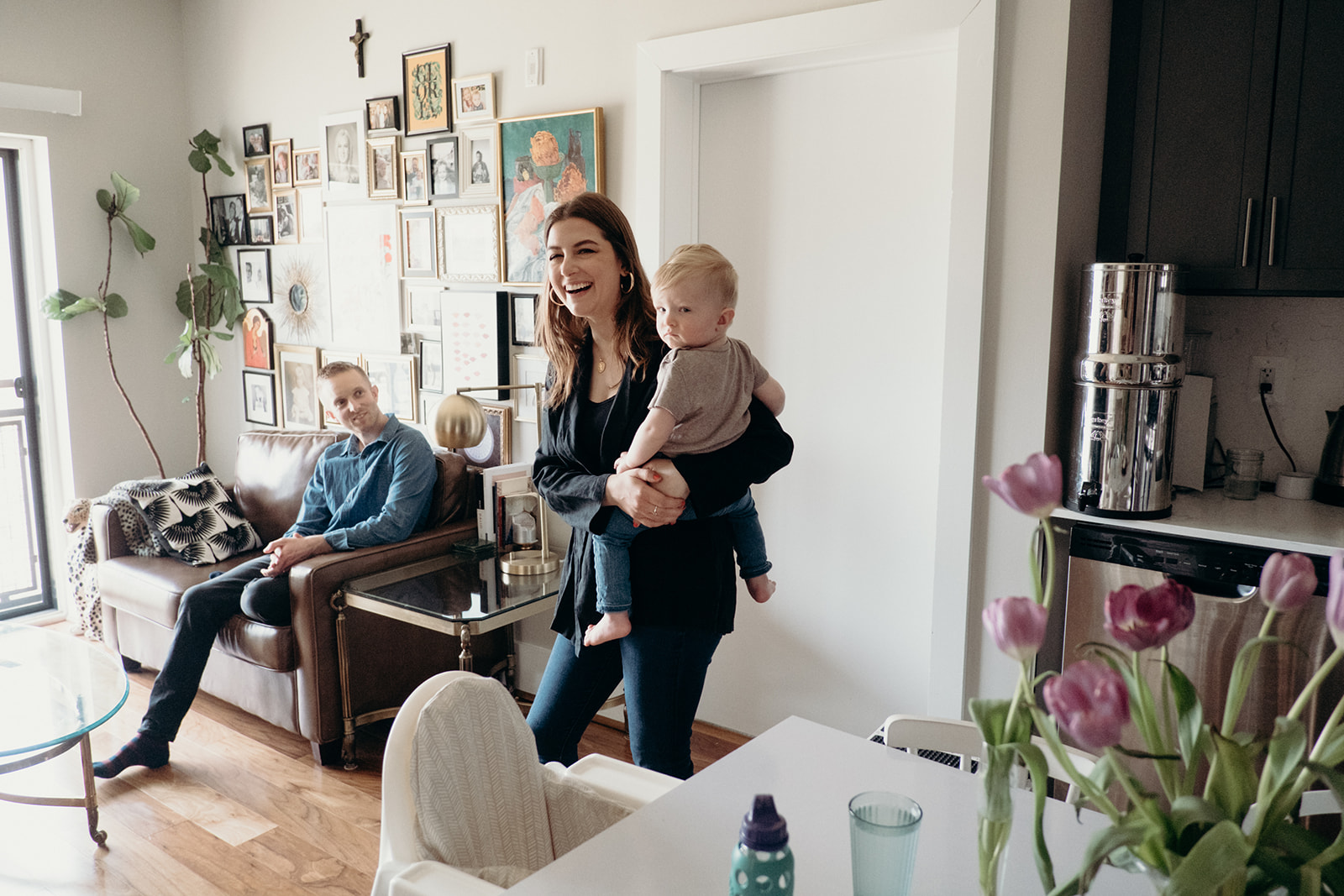 A woman holds her infant son and laughs while her husband looks on in their apartment.