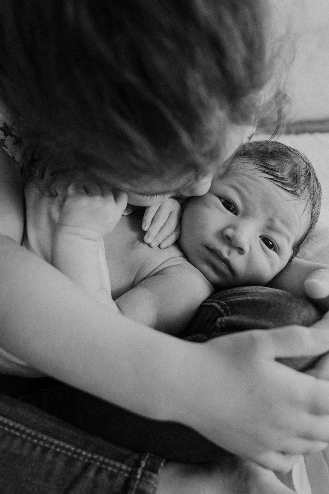 A little girl kisses her newborn baby brother while he opens his eyes during an in home family photography session. 