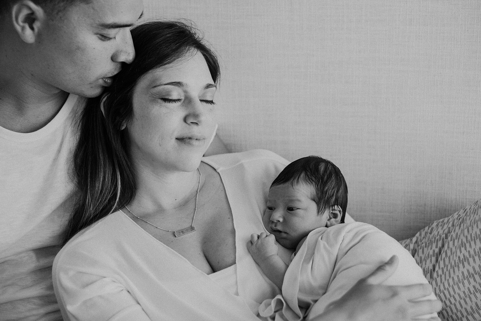 A husband holds his wife while she holds their newborn son during and in home family photography session. 