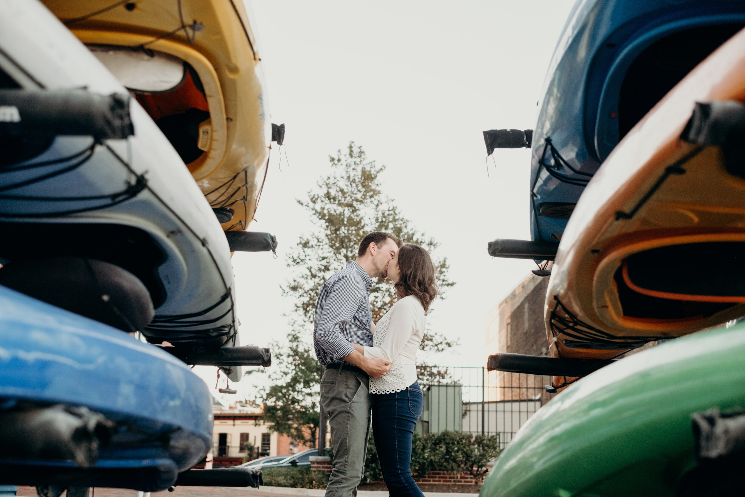 An engaged couple kisses between rows of colorful kayaks in Baltimore, MD.