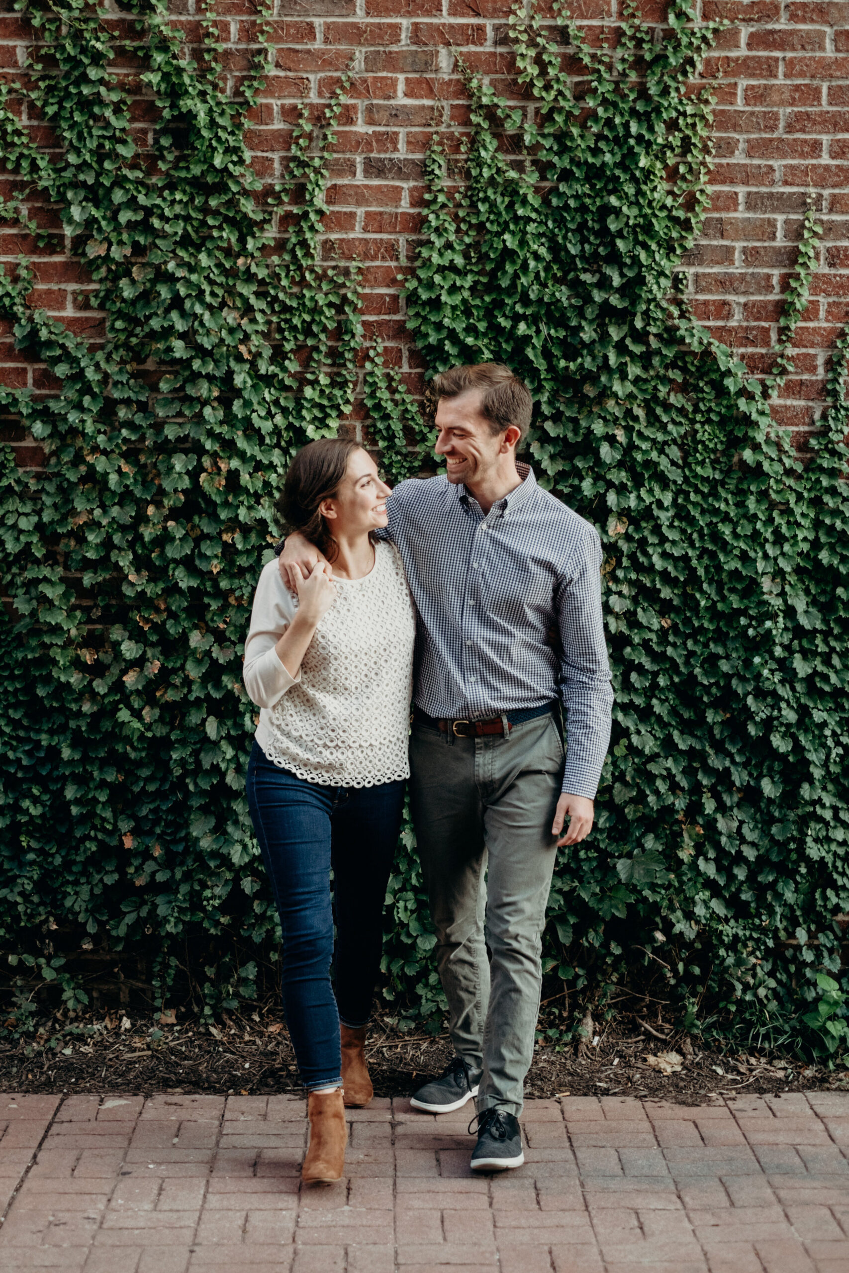 An engaged couple walks in front of a brick wall covered in ivy in Baltimore, MD.