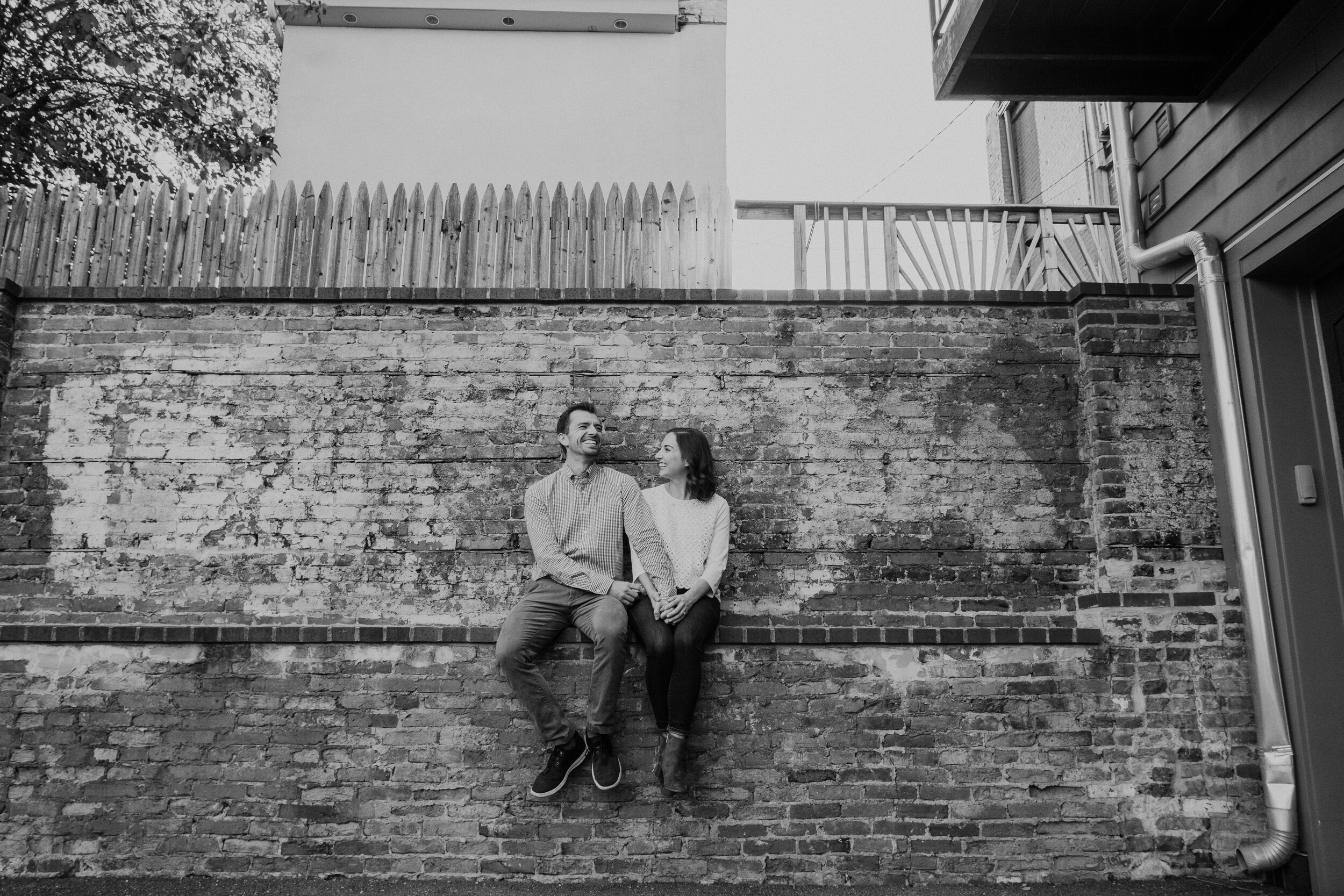 An engaged couple sits and laughs with one another on a brick wall in Baltimore, MD.