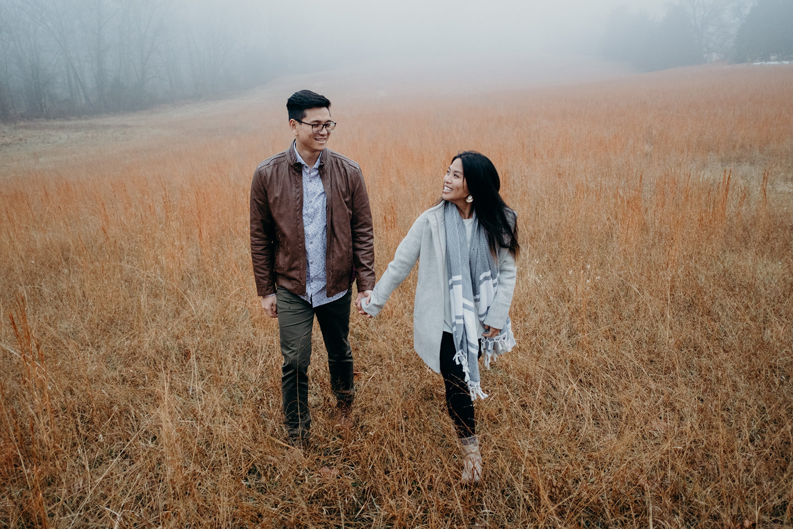 A woman smiles at her fiancé while walking with him through a grassy, foggy field at Morven Park in Leesburg, VA during their engagement photo session. 