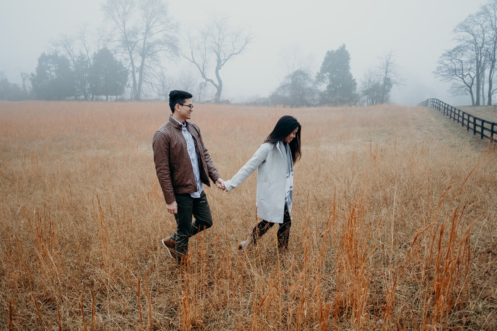 A woman leads her fiancé towards a fence surrounding a grassy, foggy field in Morven Park in Leesburg, VA during their engagement photo session.