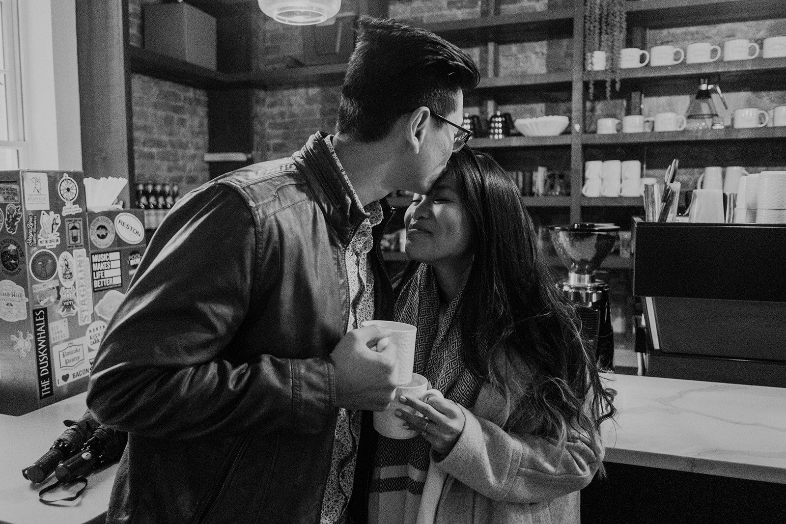 A man kisses his fiancé on the forehead while they share a coffee at SideBar coffee in downtown Leesburg, VA during their engagement photo session. 