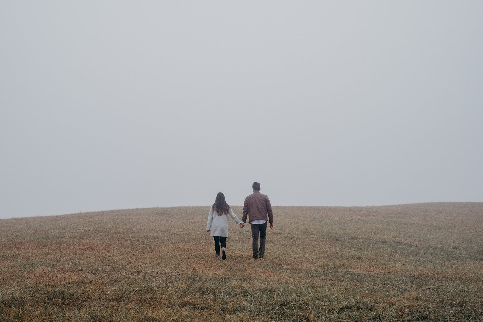 A couple climbs a foggy hill in a field at Morven Park in Leesburg, VA during their engagement photo session. 