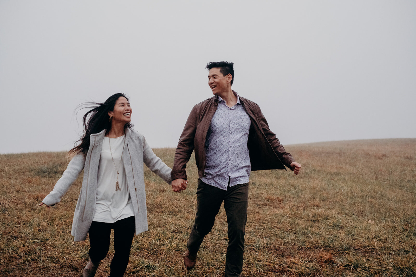 The wind whips the long hair of a woman as she runs down a hill laughing with her fiancé in Morven Park in Leesburg, VA during their engagement photo session. 