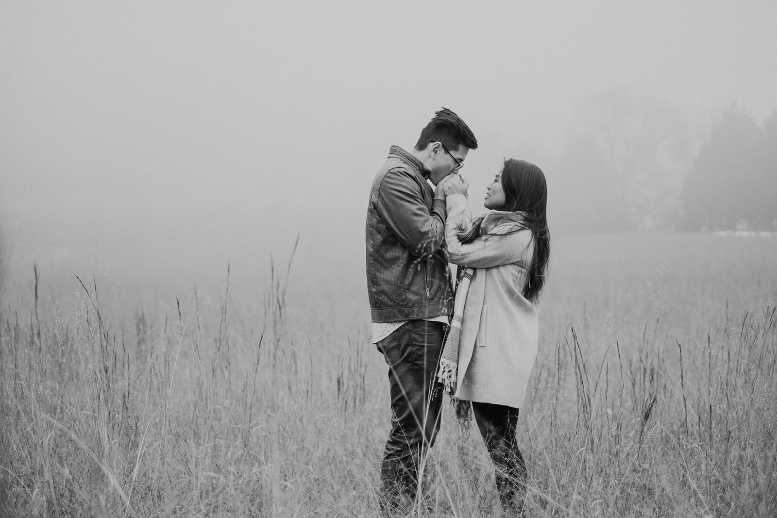 A man kisses his fiancé's hands as they stand in a grassy, foggy field in Morven Park in Leesburg, VA during their engagement photo session. 