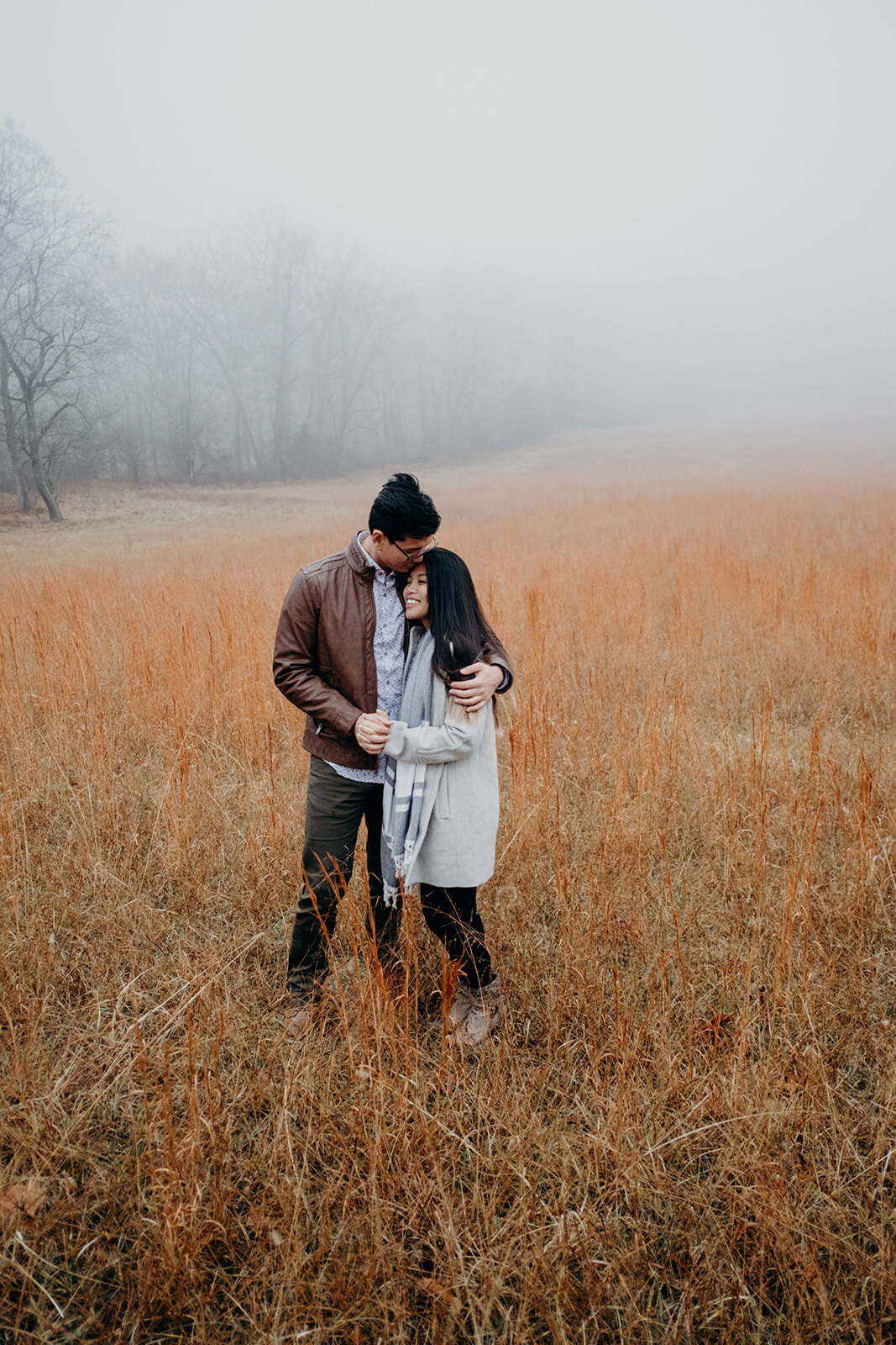 A man kisses and holds his fiancé while they stand in a grassy, foggy field in Morven Park in Leesburg, VA during their engagement photo session.