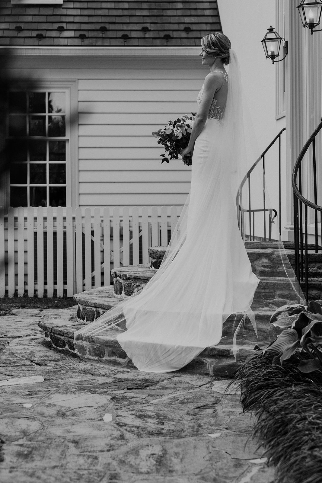 A bride in a beaded and satin wedding dress and cathedral length veil stands on the the steps of the house at the Retreat at Eastwood Farm in Warrenton, VA. 