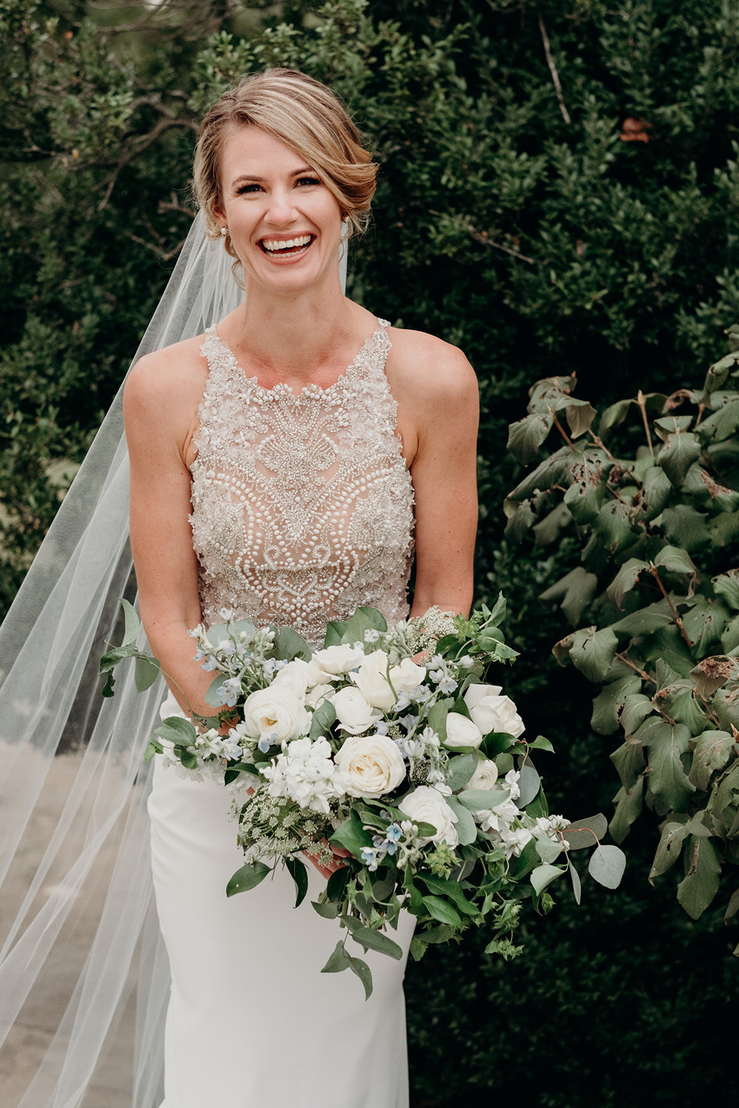 A bride carrying a bouquet of white flowers and greenery in a beaded satin dress and cathedral veil laughs on her wedding day at the Retreat at Eastwood Farm in Warrenton, VA.