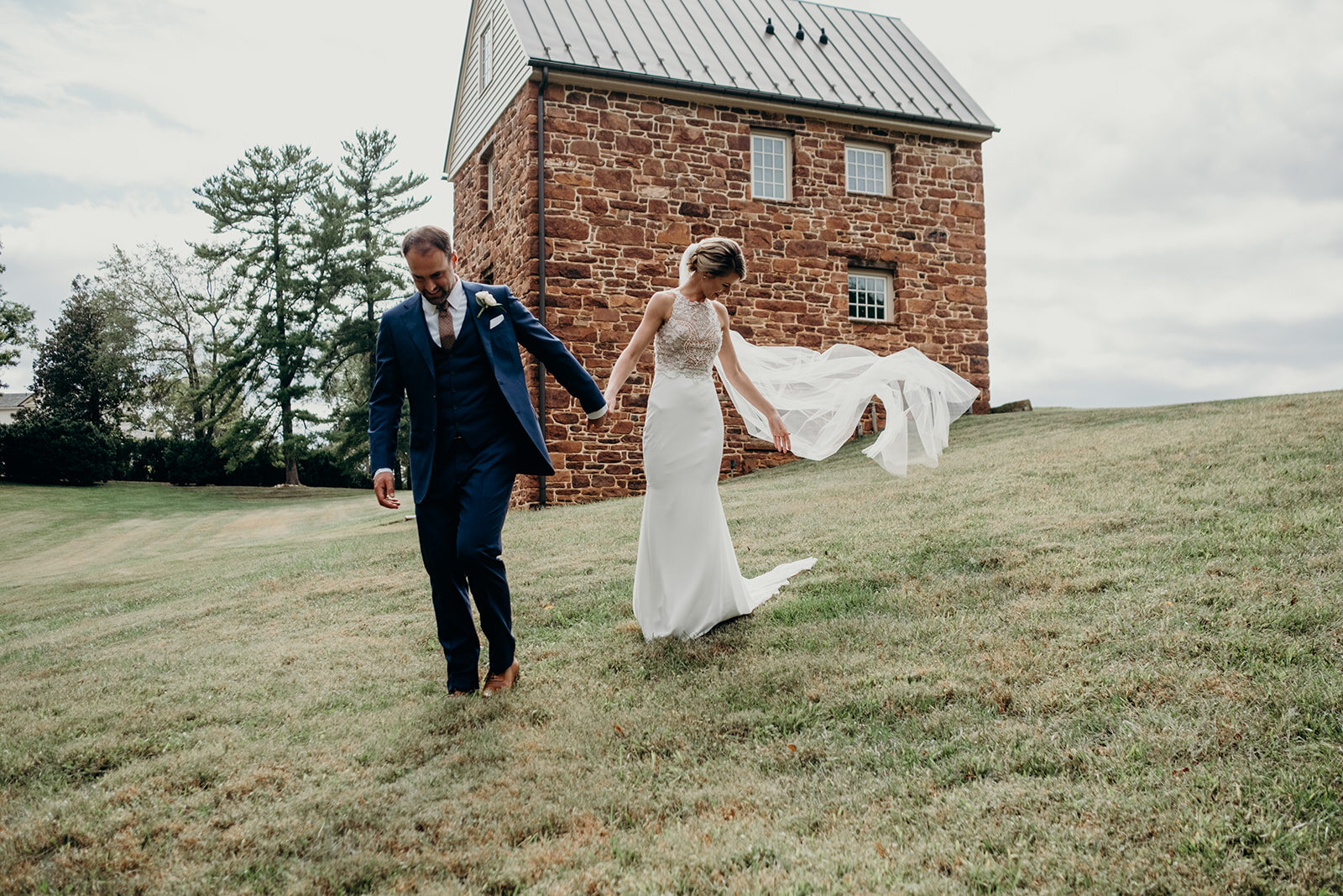 A bride and groom walk down a hill while the bride's cathedral veil trails in the wind at the Retreat at Eastwood Farm in Warrenton, VA.