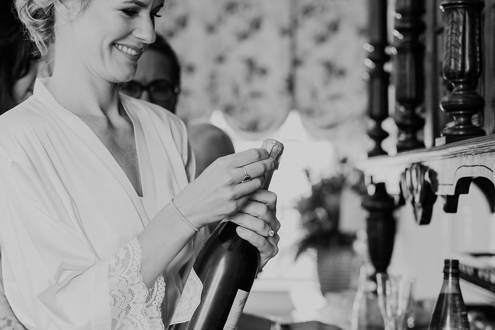 A bride in a robe opens a bottle of champagne to toast with her mother and sister before her outdoor garden wedding at the Retreat at Eastwood Farm in Warrenton, VA. 