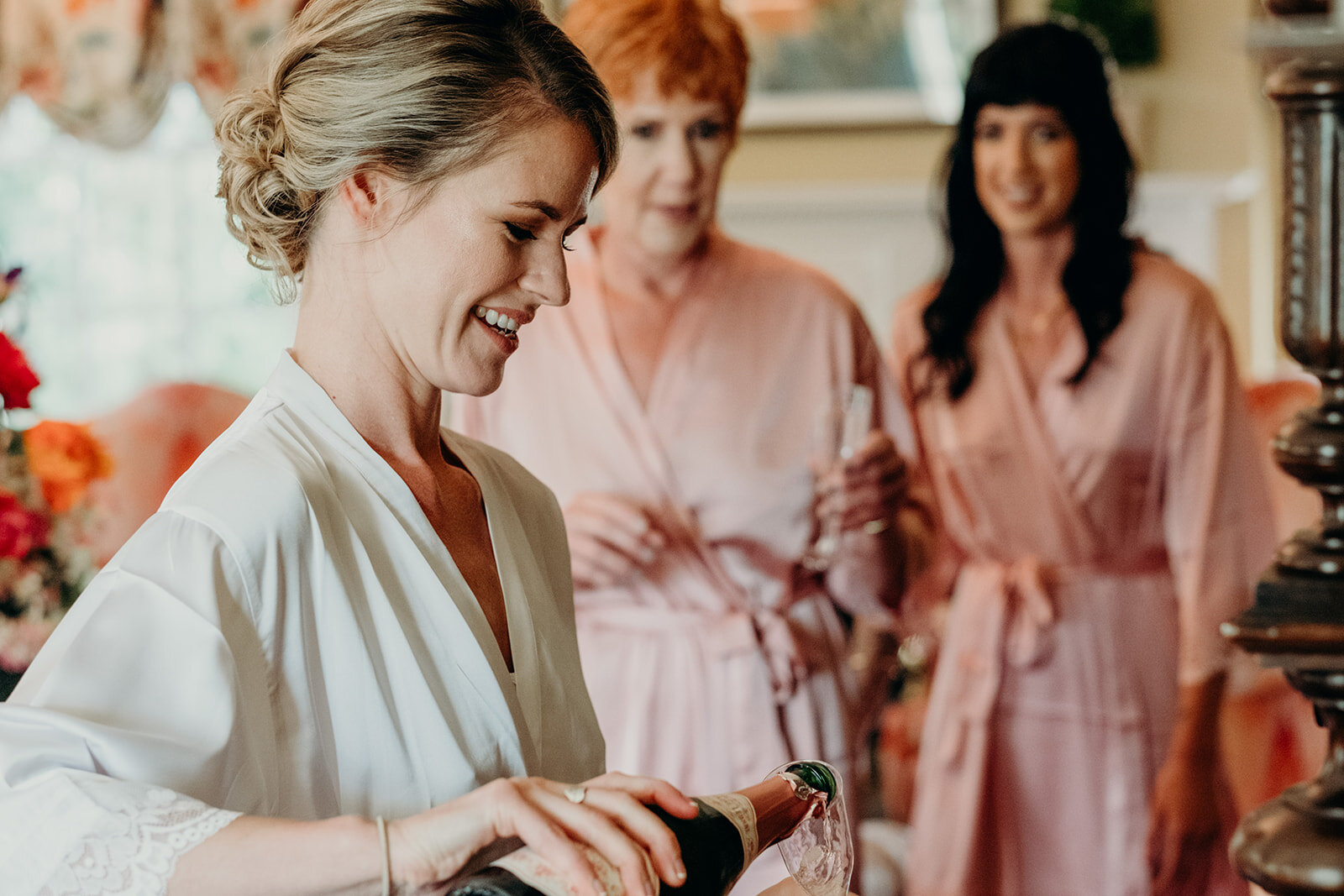 A bride in a robe pours champagne for her mother and sister before her outdoor garden wedding at the Retreat at Eastwood Farm in Warrenton, VA. 